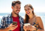 Aussie couple laughing, holding phones, beach backdrop.